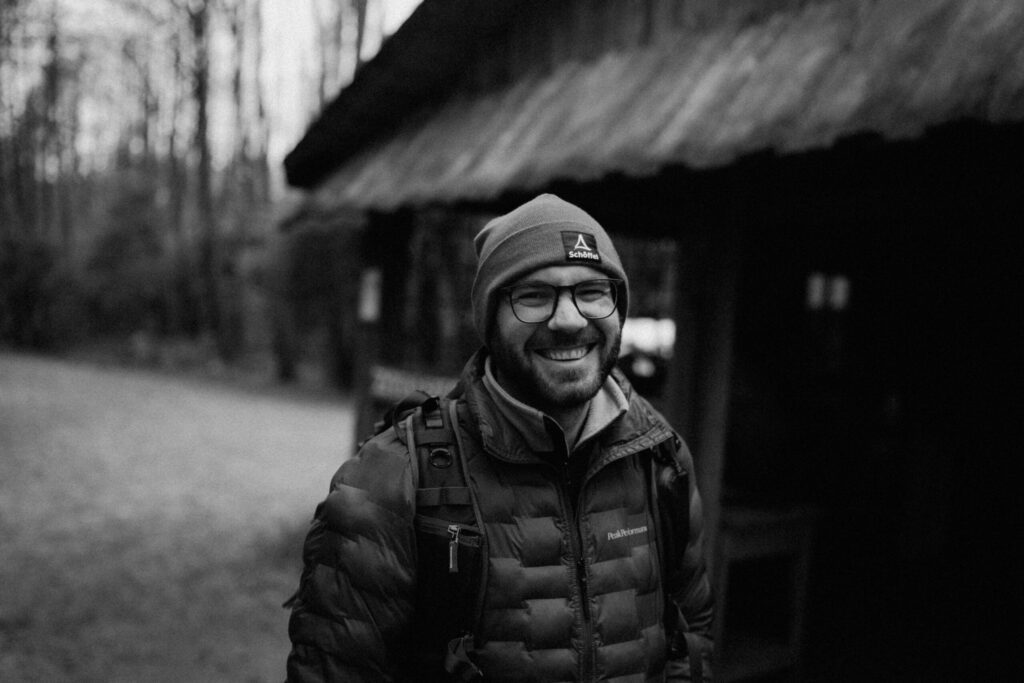 Ein Mann mit Brille, Beanie und dicker Jacke lächelt draußen. Er trägt einen Rucksack und steht neben einer rustikalen Holzhütte in einem Waldgebiet. Das Bild ist schwarzweiß.